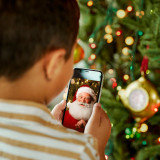 Looking over the shoulder of a young boy watching a Santa video on a phone in front of the Christmas tree.