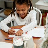 A smiling young girl drawing with crayons in a Santa Kindness journal at a holiday table.