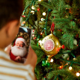 An image looking over the shoulder of a young boy who is seeing a message from Santa on a phone after scanning the Santa's Kindness ornament hanging on the Christmas tree.